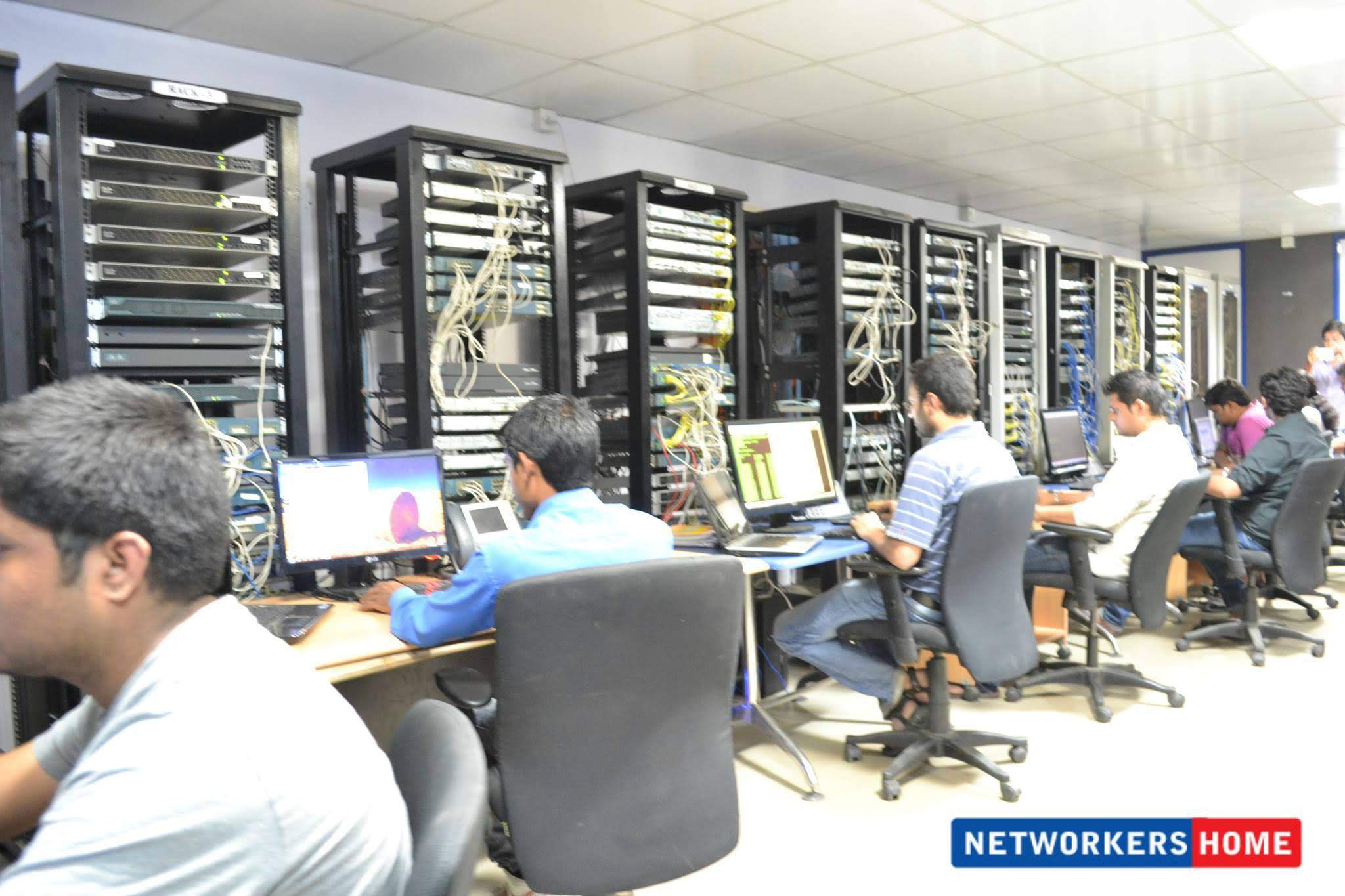 Wide view of Networkers Home multi-rack networking lab with 6+ students working on real Cisco hardware